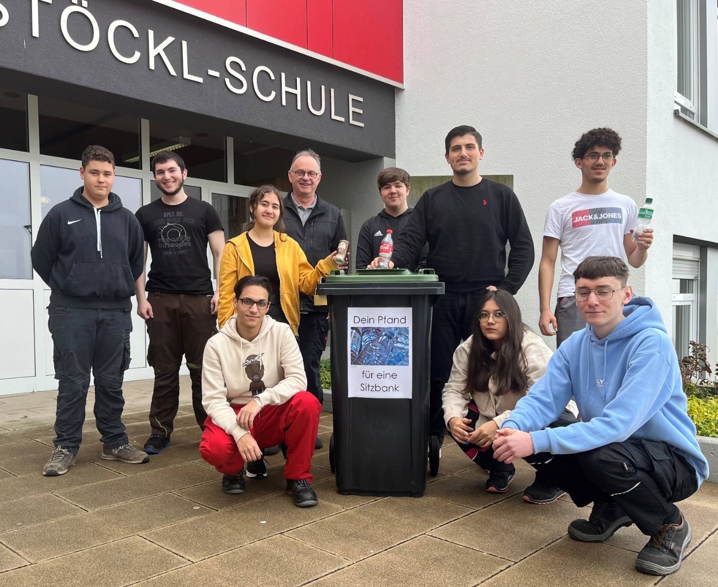 Lernende der Berufsfachschule Mechatronik mit Schulsprecher Murat Kaya (7. v.l.) und der betreuenden Lehrkraft Reinhard Schmidt an dem gefertigten Sammlungsbehälter für Pfandflaschen.